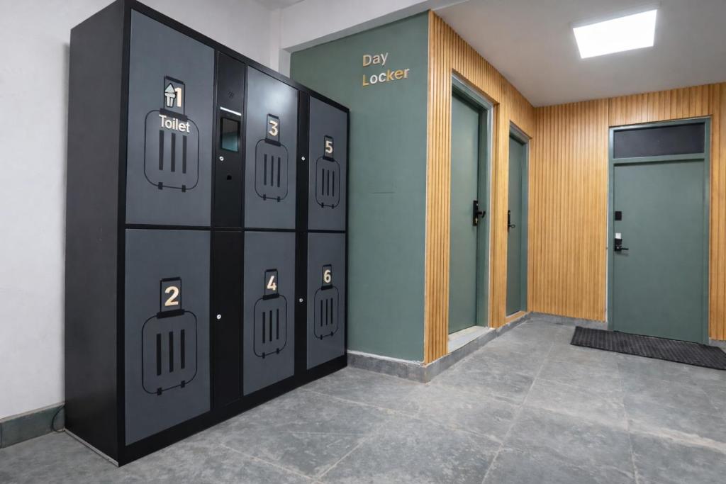 Tiny Living Apartments Day Locker — matte-black six-door locker unit with numbered doors (1 to 6, plus a 'Toilet' tile), located in a brightly lit ground-floor corridor with green doors leading to the building